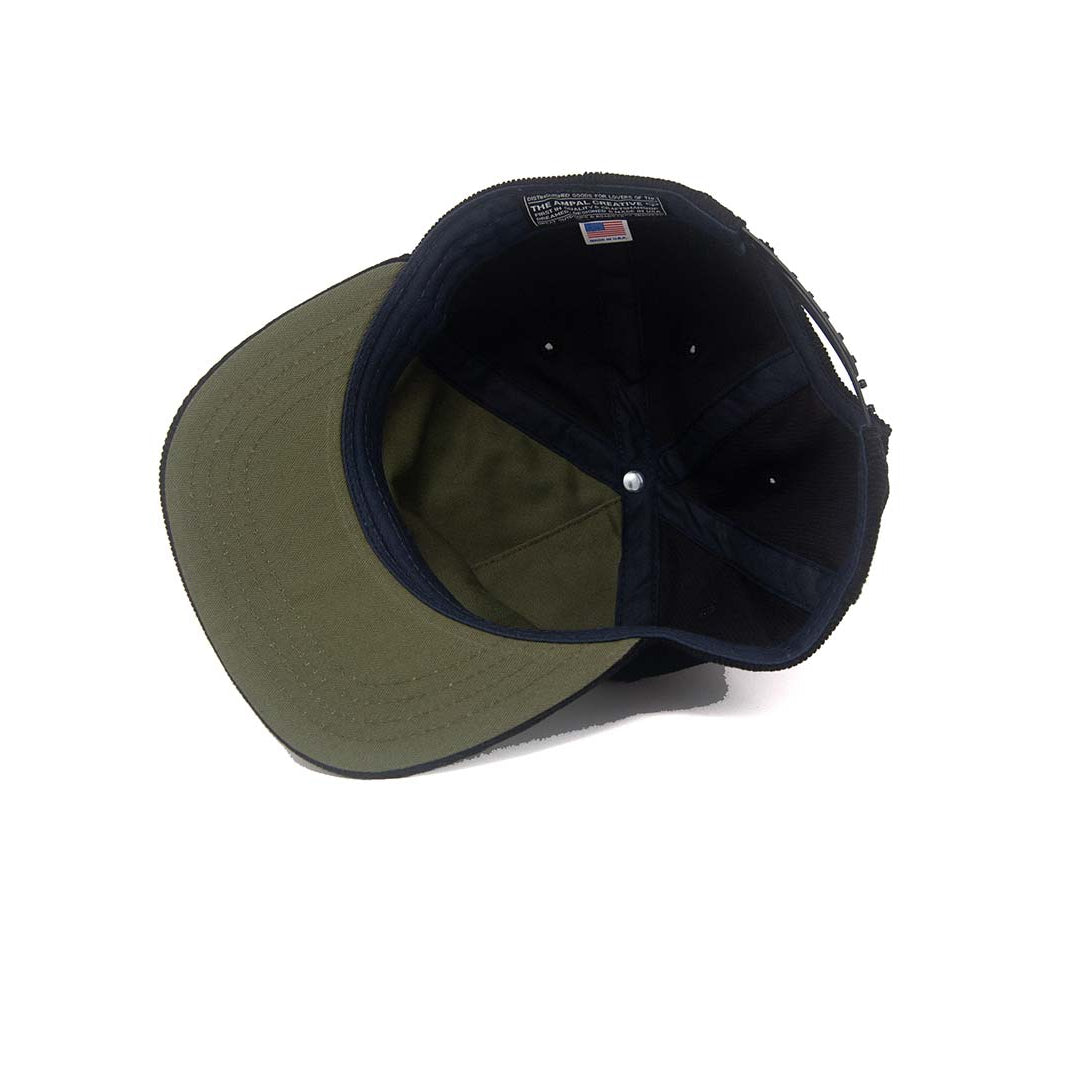 Interior view of the Ampal Creative CA Snapback in Black Corduroy, showing the olive herringbone underbrim, navy crown lining, The Ampal Creative woven label with USA flag, and snapback closure, on a white background
