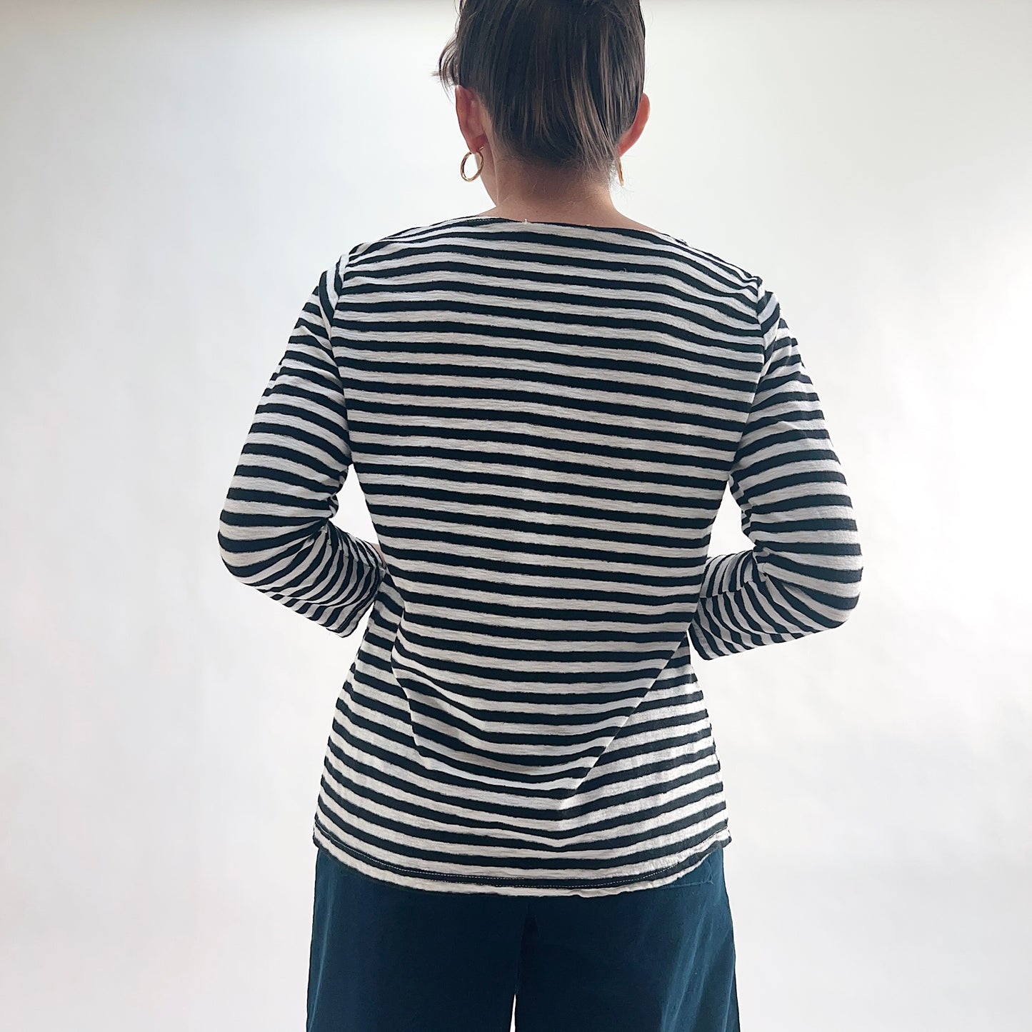 Back view of model wearing a navy and white laundered stripe 3/4 sleeve boatneck tee, hands behind back, showing the wide boatneck and curved hem, styled with dark blue wide-leg pants.
