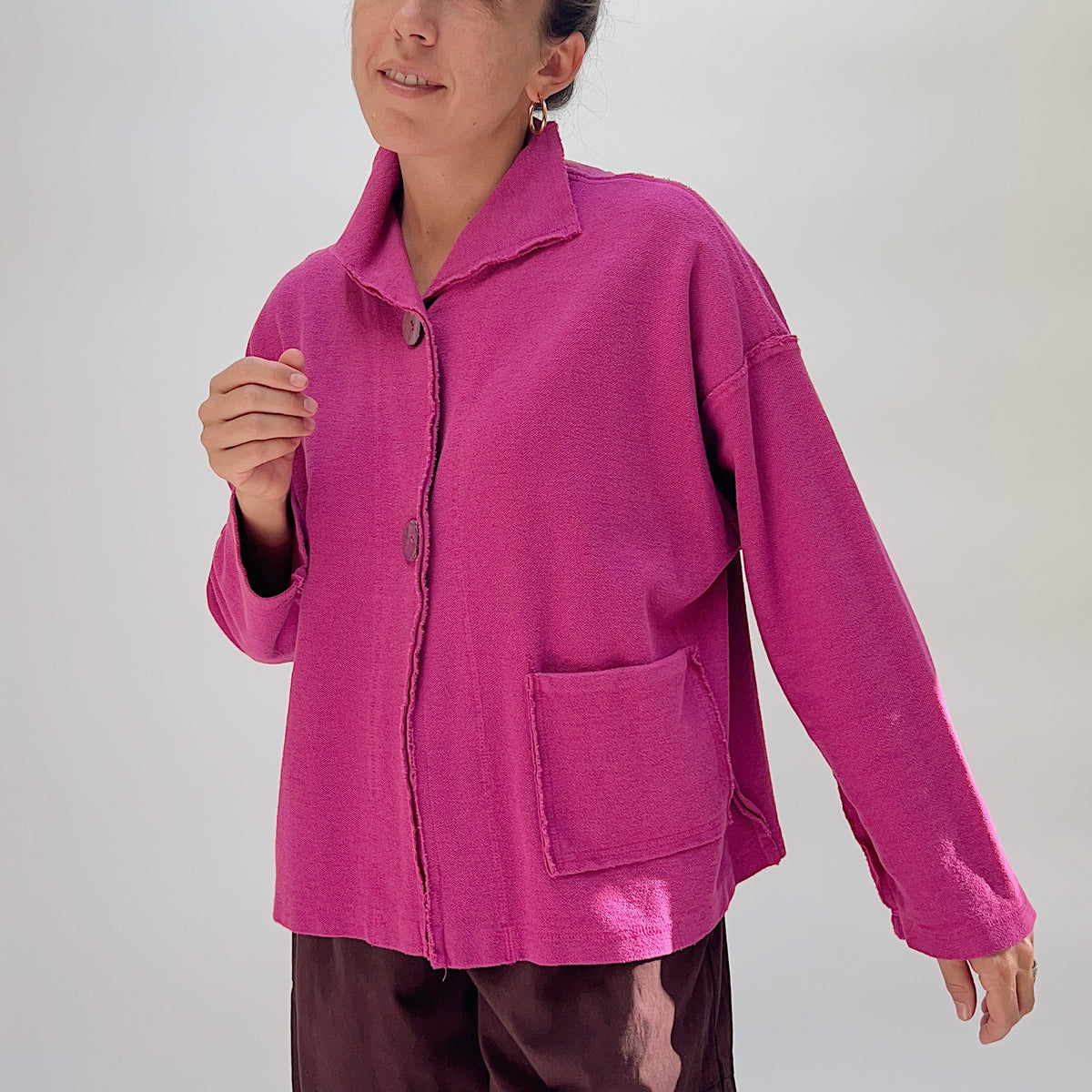 Eleven Stitch Funnel Jacket in Berry, three-quarter front view on model, berry pink 100% cotton with funnel collar, frayed seam detail, two front patch pockets, and wood buttons, paired with dark brown wide-leg trousers
