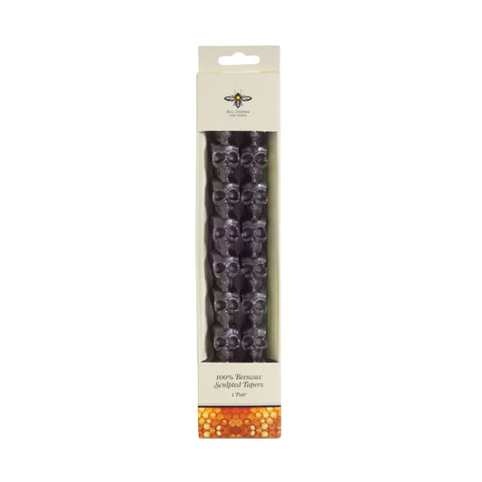 Big Dipper Wax Works 10-inch Sculpted Black Skull Tapers in retail packaging, front view against a white background, showing two dark charcoal beeswax taper candles with a stacked skull silhouette shape inside a white windowed box labeled 100% Beeswax Sculpted Tapers, 1 Pair, with a Big Dipper Wax Works logo at the top and a honeycomb pattern at the base
