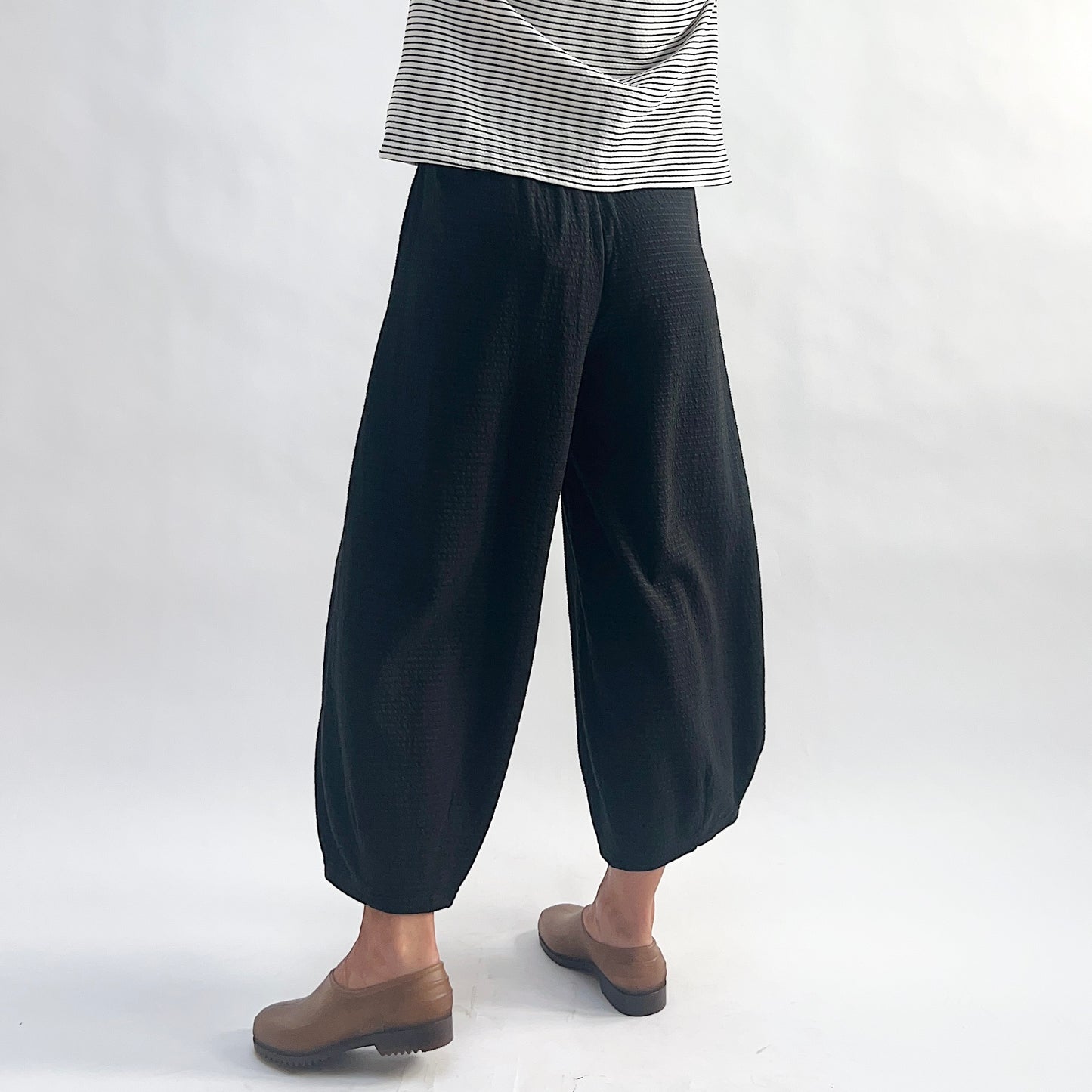 Three-quarter back view of model wearing black crinkle-texture cropped wide-leg pants, showing the elastic waist, wide rounded volume, and tapered ankle, styled with a white and black stripe long-sleeve top and taupe slip-on shoes against a white background.
