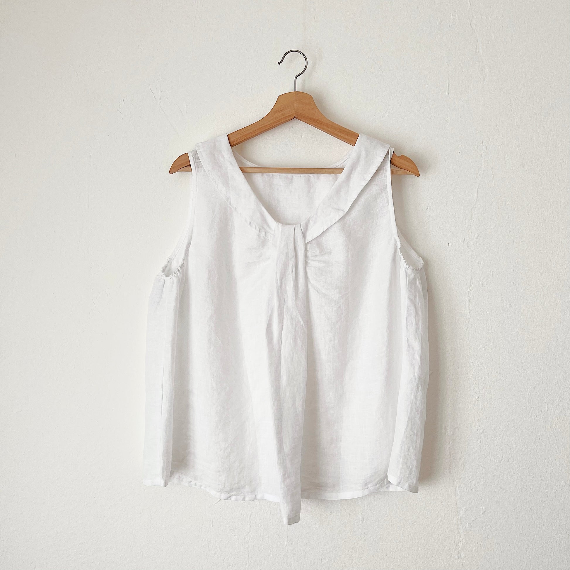 A white linen sleeveless top with a round neckline, hanging on a wooden hanger against a white wall.
