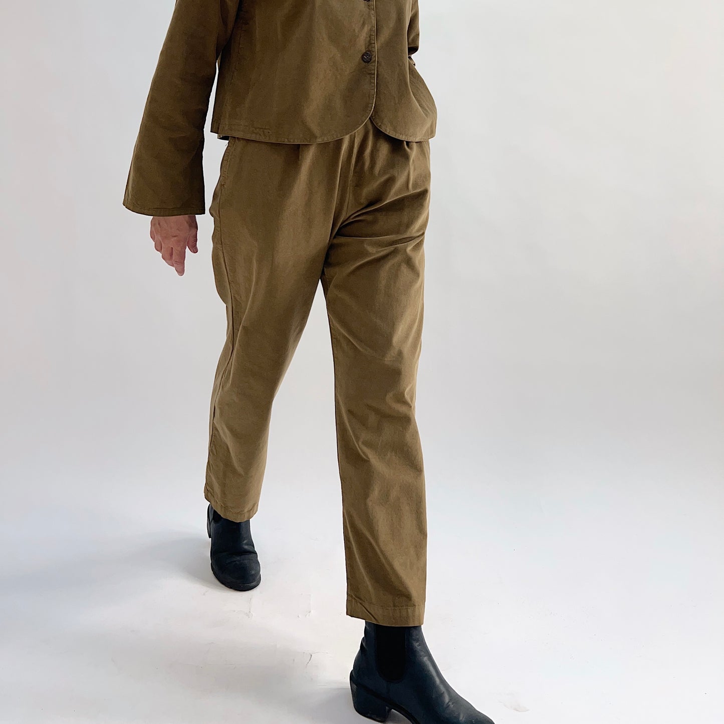 Model in motion wearing the Bryn Walker Corduroy Pleated Pants in Partridge, cropped front view showing front pleats, straight leg silhouette, and olive pinwheel corduroy, styled with matching Standing Collar Crop Jacket and black Chelsea boots
