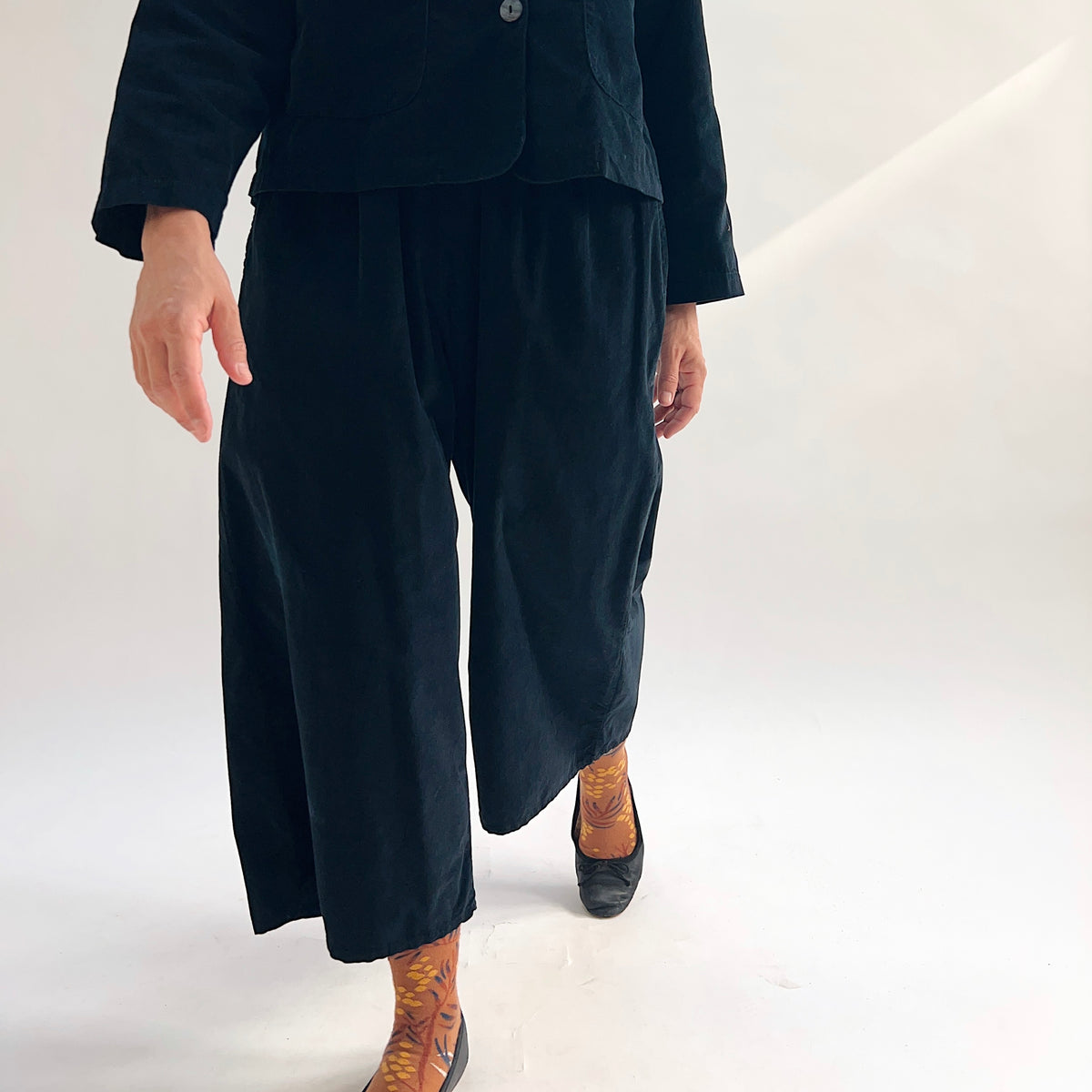 Model in motion wearing the Bryn Walker Corduroy Flood Pants in Black, cropped front view showing wide-leg silhouette, elastic waistband, and flood-length hem in black pinwheel corduroy, paired with matching jacket, printed socks, and black wedge boots
