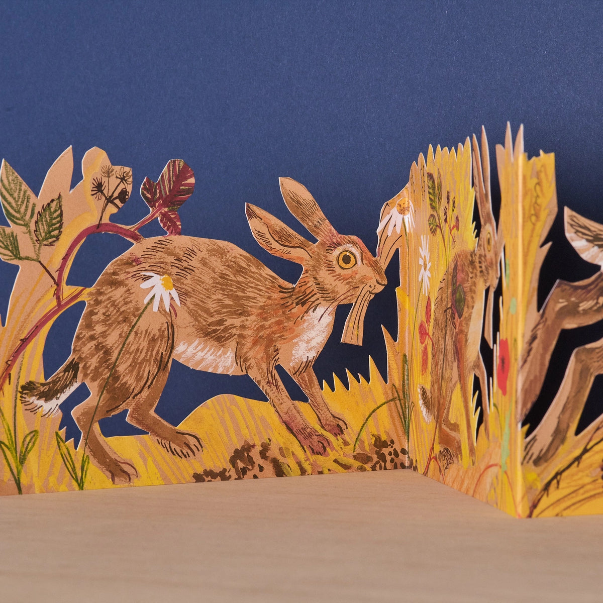 Close-up detail of the Hares Collage concertina card showing an illustrated hare leaping through golden wheat stalks and botanical foliage