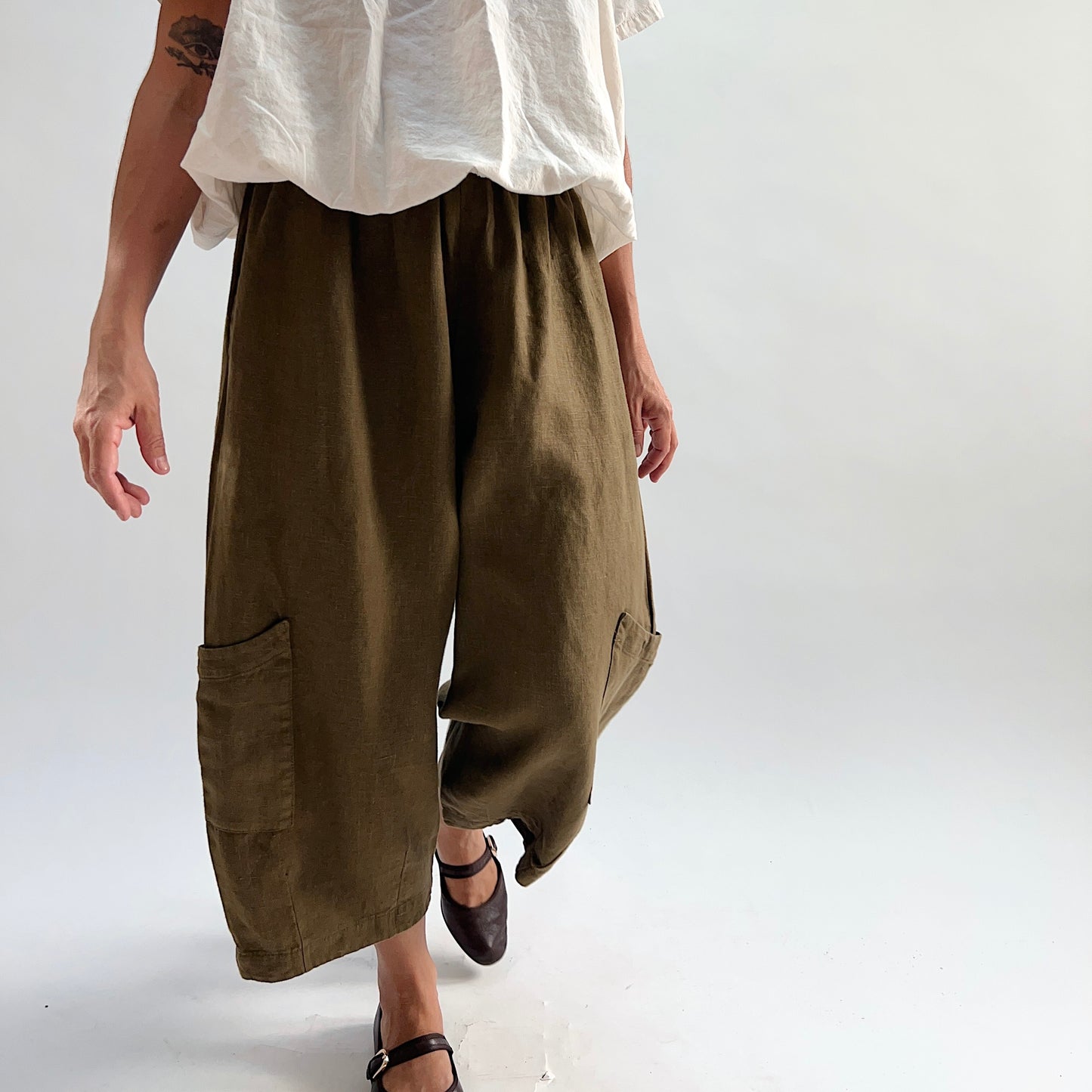 Model in motion wearing the Bryn Walker Heavy Linen Casbah Pant in Partridge, angled front view showing patch pockets, wide-leg silhouette, and olive heavyweight linen, paired with white linen top and dark Mary Jane flats
