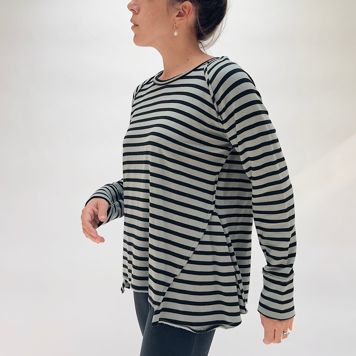Side profile of model wearing the Cut Loose Striped Gore Tee in Seal, showing the angled gore panels at the sides and hem that create an A-line swing silhouette, raglan seams, thumb-hole cuffs, and raw lettuce-edge hem in seal grey and black horizontal stripe tencel-cotton blend
