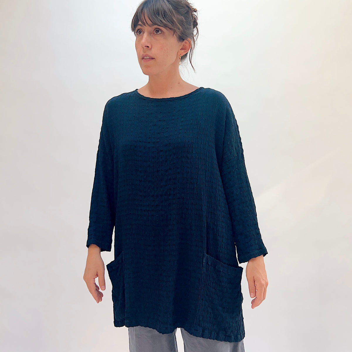 Three-quarter side view of teal crinkle-texture one-size pocket tunic showing scoop neckline, 3/4 sleeves, and side patch pocket detail, styled with grey wide-leg pants, gold drop earrings
