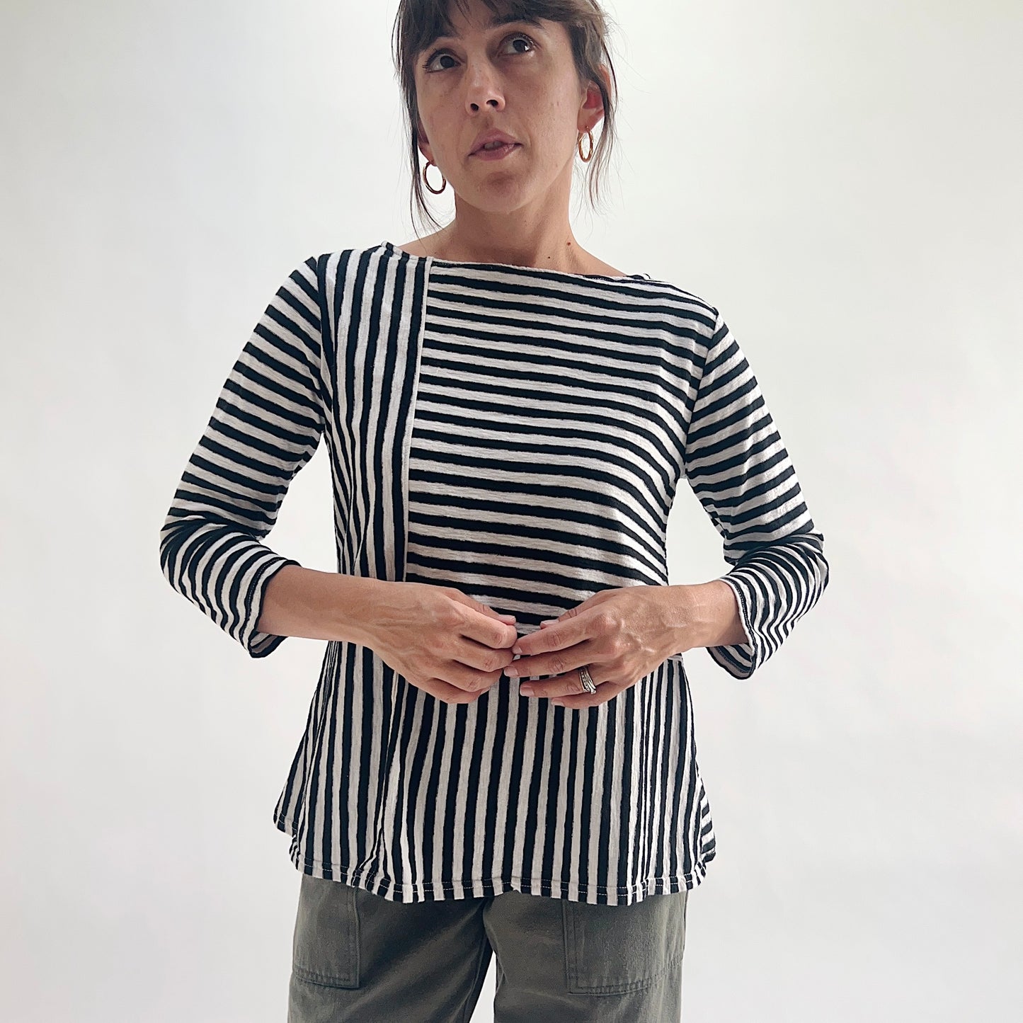 Front view of model wearing a black and white patchwork stripe A-line boatneck top with 3/4 sleeves, featuring alternating vertical and horizontal stripe panels, styled with olive green pants against a white background.
