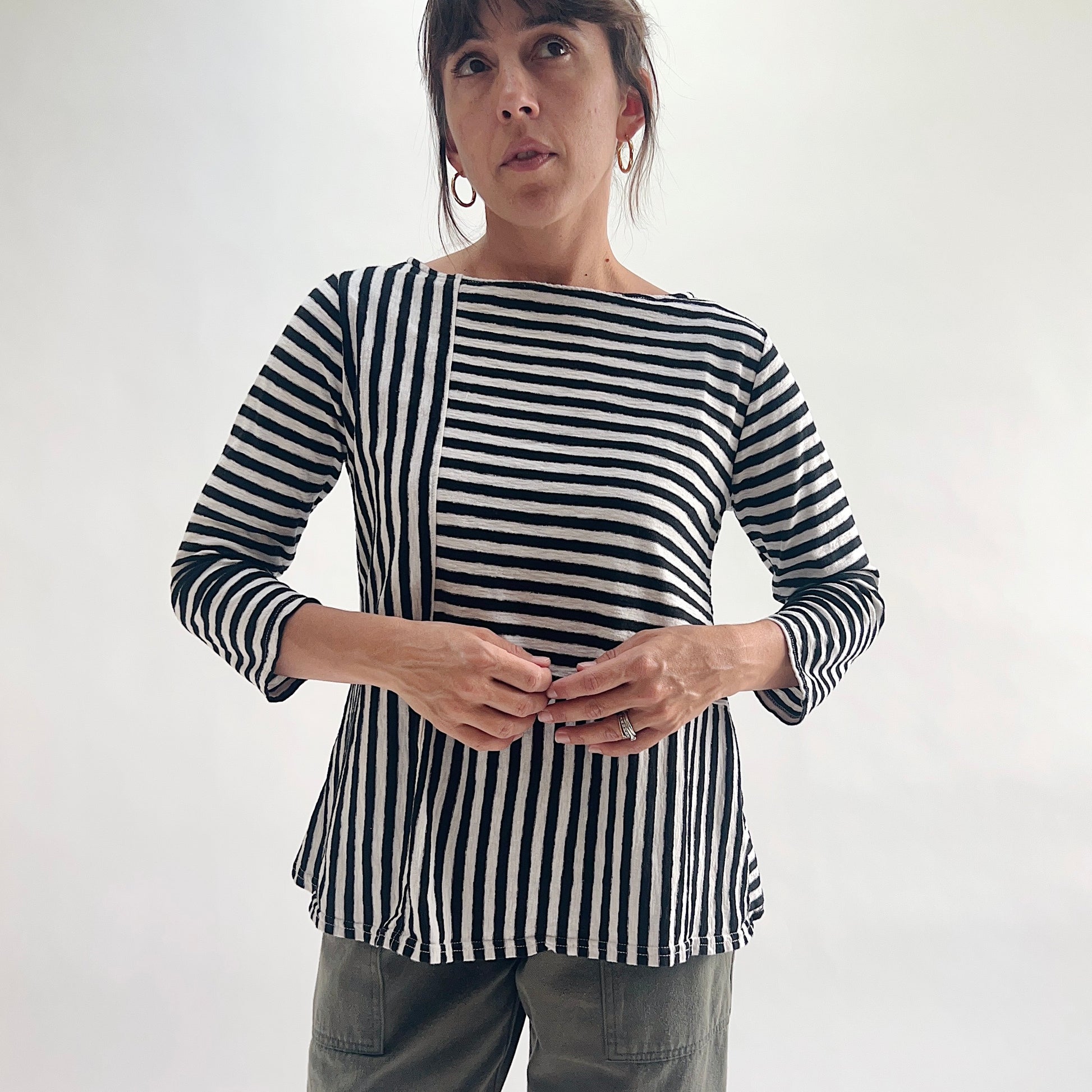 Front view of model wearing a black and white patchwork stripe A-line boatneck top with 3/4 sleeves, featuring alternating vertical and horizontal stripe panels, styled with olive green pants against a white background.
