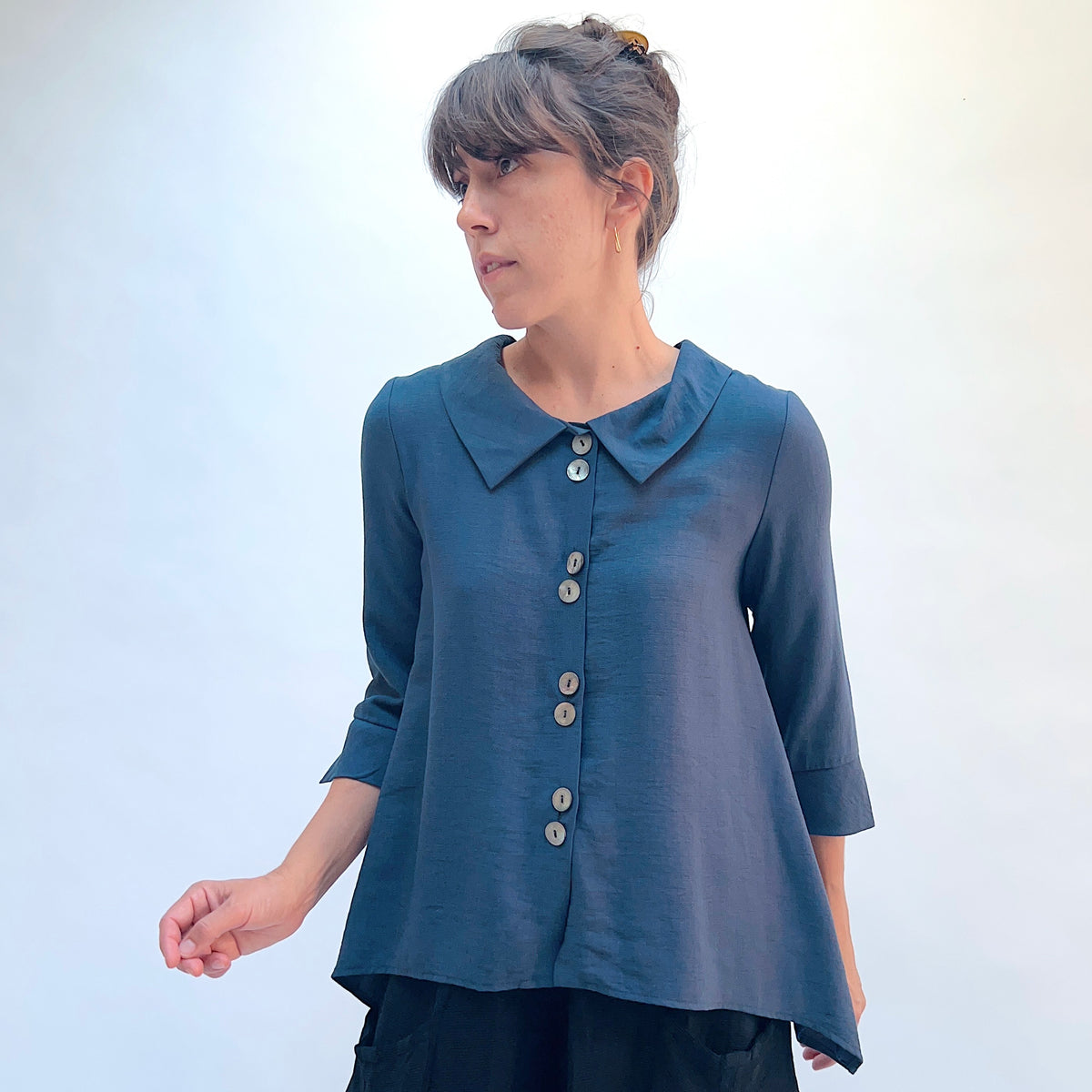 A woman wearing a slate blue button-up blouse with a pointed collar and three-quarter sleeves, paired with black pants, set against a plain light background.