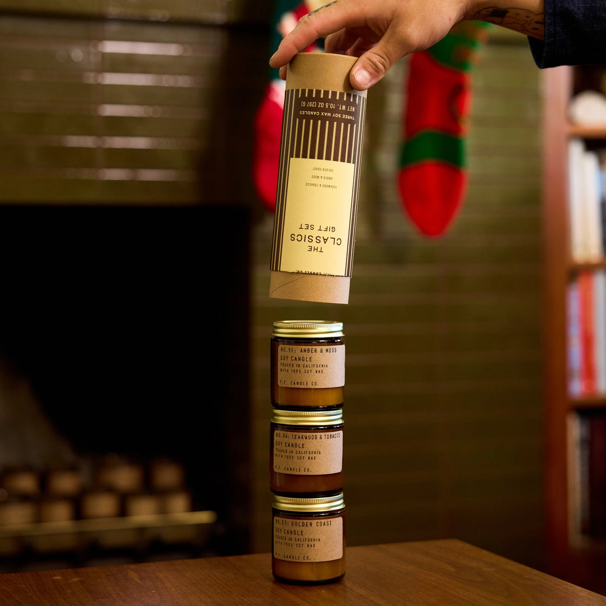 A hand lifts The Classics gift tube above three stacked mini candles: Amber & Moss, Teakwood & Tobacco, and Golden Coast.
