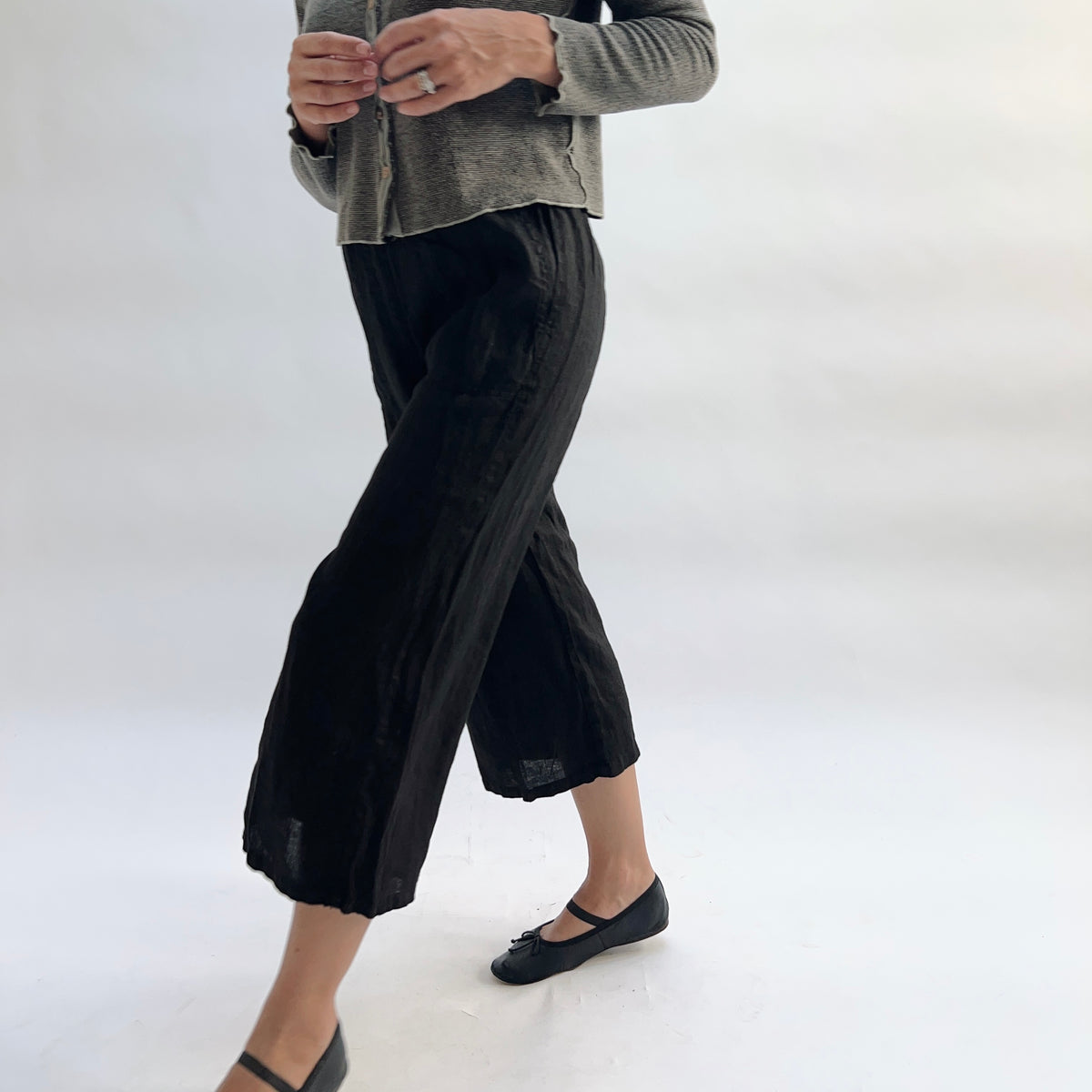Cut Loose Hanky Linen Easy Crop Flood Pant in Black, black linen wide-leg crop pant mid-stride with legs crossed, styled with a grey button-front cardigan and black Mary Jane flats — side view showing drape, linen texture, and hem vent

