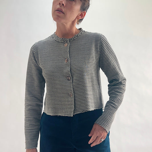 Front view of model wearing a squirrel and black fine horizontal stripe cropped cardigan with asymmetrical button closure, band neckline, and long sleeves, hand on hip, showing the crinkle-texture fabric and cropped length, styled with dark teal wide-leg pants against a white background.
