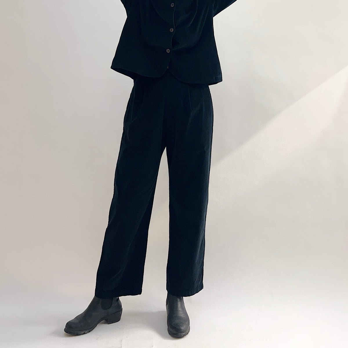 Model wearing the Bryn Walker Corduroy Pleated Pants in Black, full-length front view highlighting front pleats, straight leg, full-length hem, and black pinwheel corduroy texture, styled with matching Standing Collar Crop Jacket and black Chelsea boots
