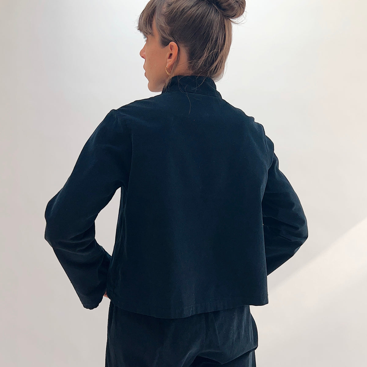 Back view of model wearing the Bryn Walker Corduroy Standing Collar Crop Jacket in Black, showing ruffled standing collar, wide sleeves, clean cropped hemline, and black pinwheel corduroy texture, paired with matching pants
