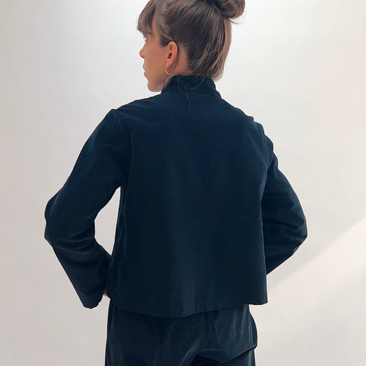 Back view of model wearing the Bryn Walker Corduroy Standing Collar Crop Jacket in Black, showing ruffled standing collar, wide sleeves, clean cropped hemline, and black pinwheel corduroy texture, paired with matching pants

