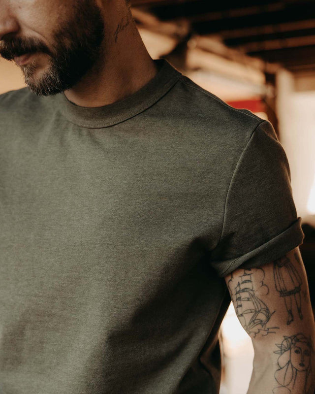 Close-up of man wearing the Taylor Stitch Organic Cotton Shop Tee in Army Twist, showing ribbed crewneck, shoulder seam detail, and cuffed sleeve on heathered army green cotton jersey
