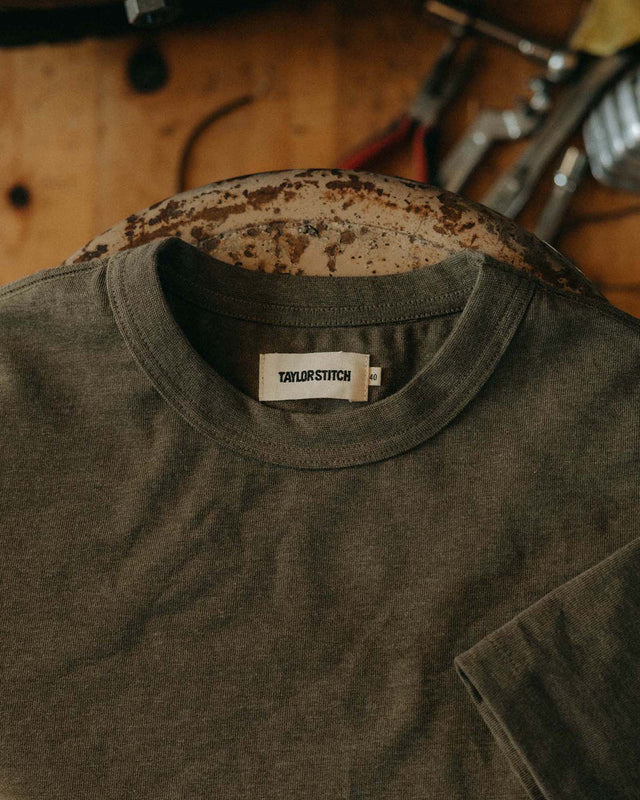 Taylor Stitch Organic Cotton Shop Tee in Army Twist on a hanger in a workshop setting, showing ribbed crewneck and Taylor Stitch woven label on heathered army green cotton
