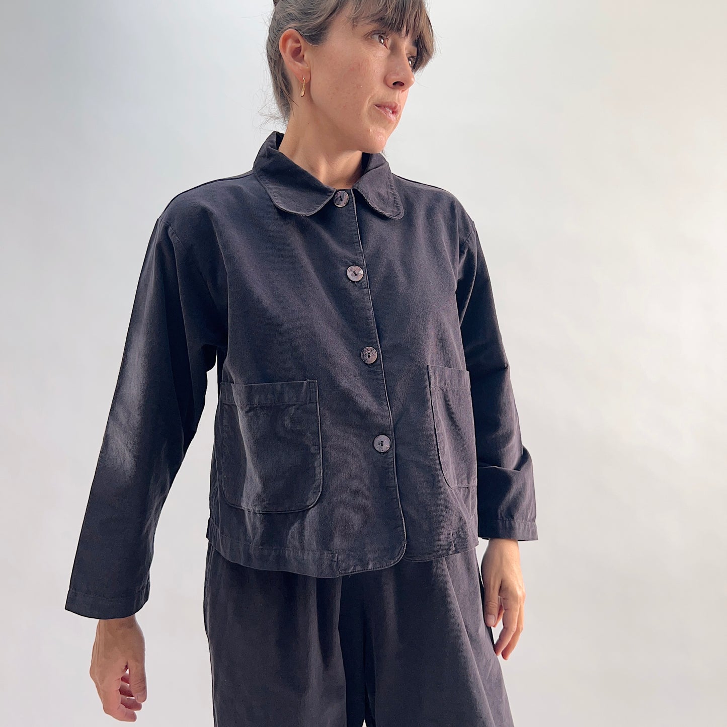 Model wearing the Bryn Walker Corduroy Audrey Jacket in Fig, angled front view showing rounded peter-pan collar, shell buttons, and patch pockets, paired with matching charcoal trousers
