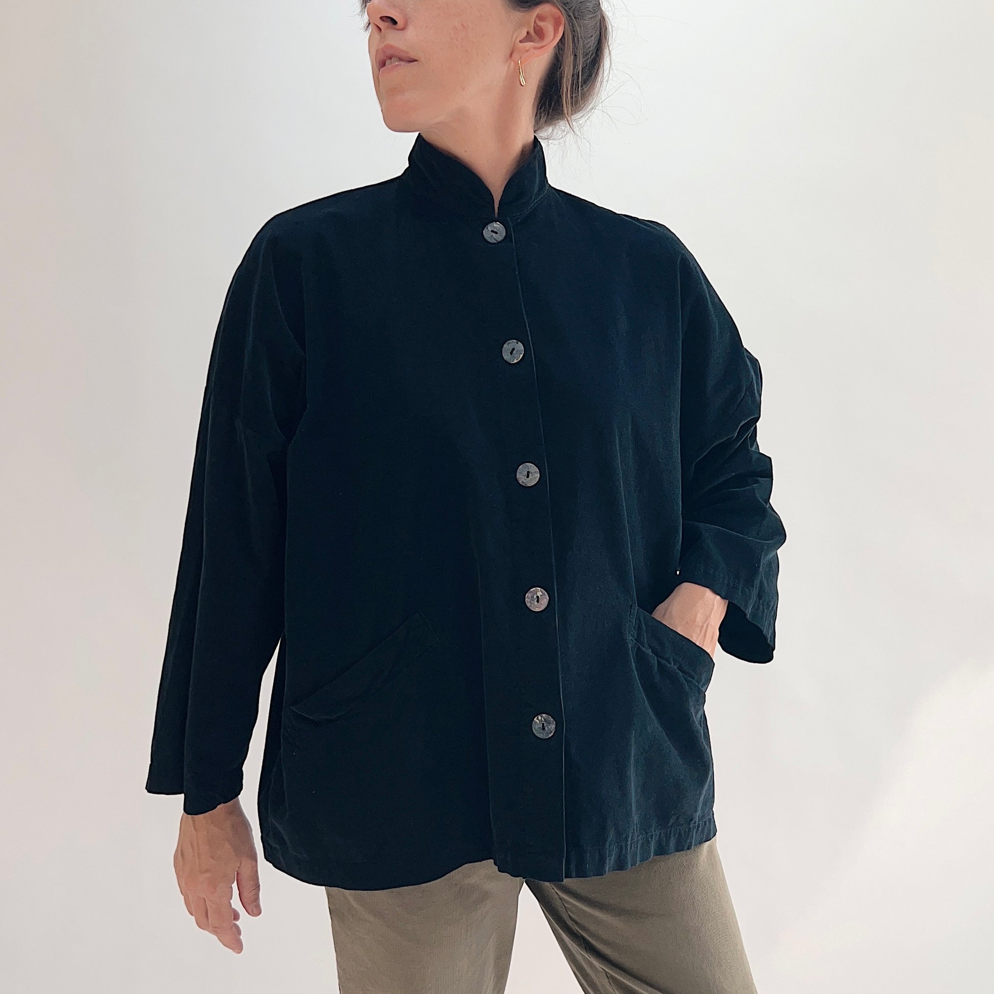Model with hand in pocket wearing the Bryn Walker Corduroy Danuta Jacket in Black, full front view showing standing collar, shell buttons, wide dolman sleeves, front patch pockets, curved hem, and black pinwheel corduroy, styled with olive pants
