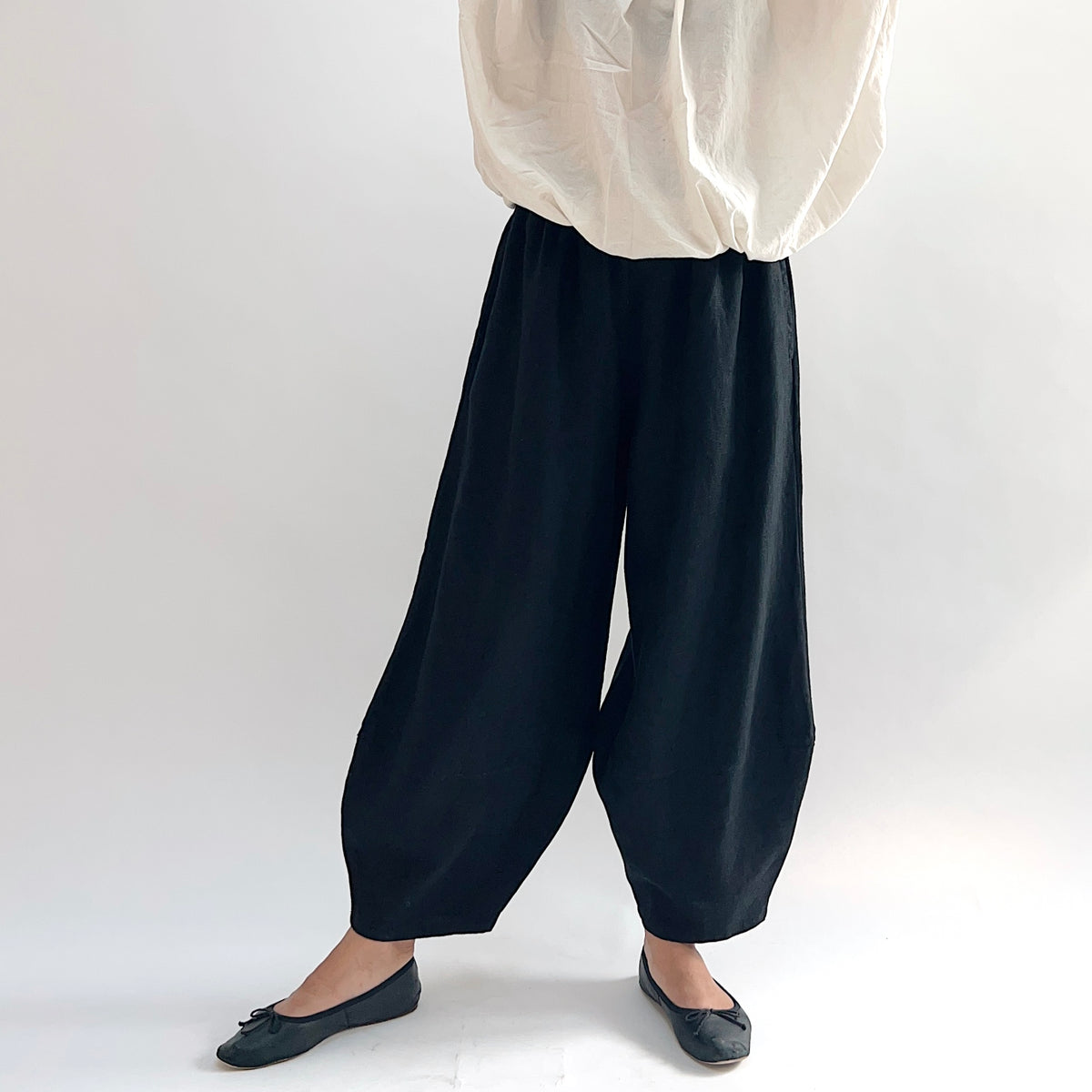 Model wearing the Bryn Walker Heavy Linen Oliver Pant in Black, angled front view highlighting barrel-leg cocoon drape, elastic waistband, and tapered ankle in black heavy linen, styled with cream long-sleeve linen top and black ballet flats

