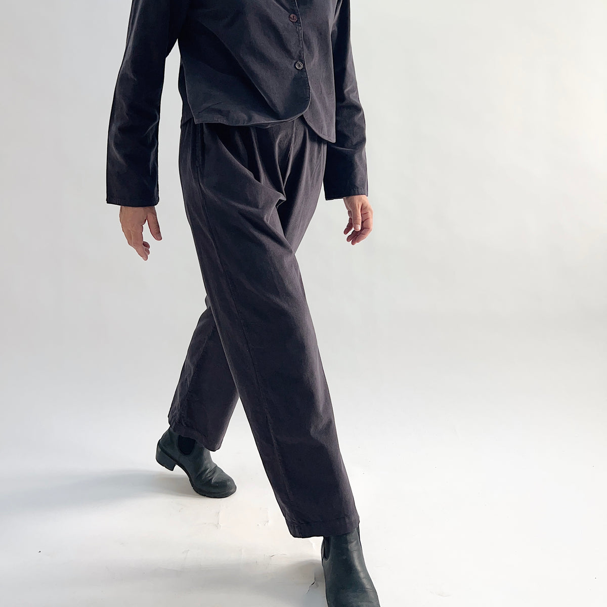 Model walking in the Bryn Walker Corduroy Pleated Pants in Fig, showing movement in tapered straight leg silhouette, flat front with pleats, and deep fig pinwheel corduroy, styled with matching Standing Collar Crop Jacket and black Chelsea boots

