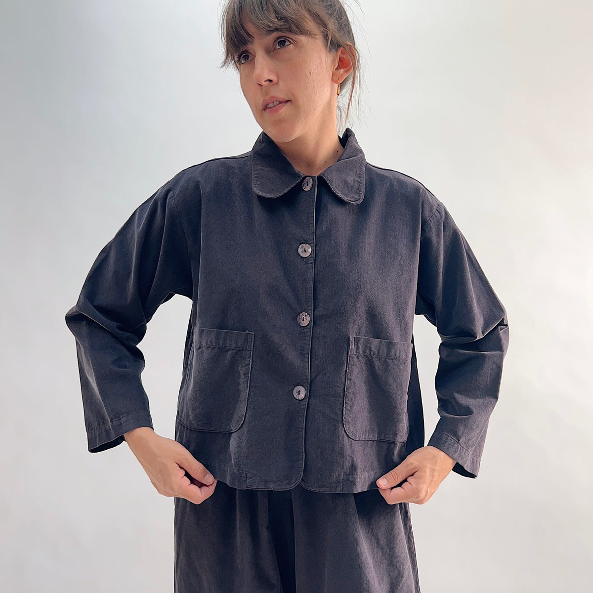 Model with hands on hips wearing the Bryn Walker Corduroy Audrey Jacket in Fig, full front view highlighting rounded collar, shell button closure, patch pockets, and deep charcoal pinwheel corduroy
