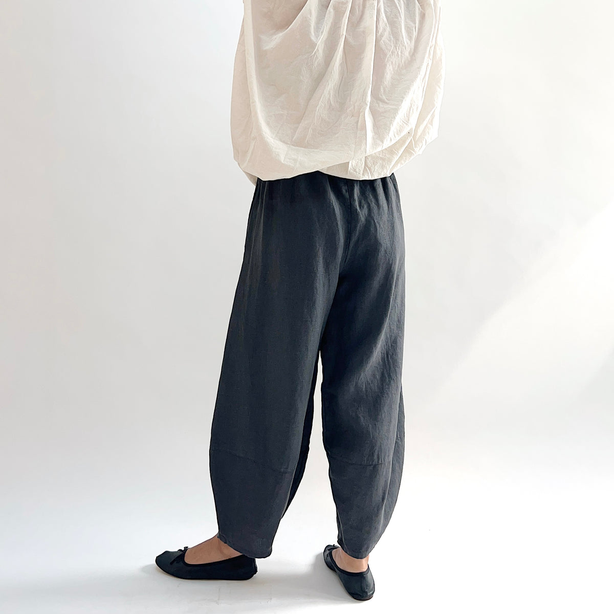 Back view of model wearing the Bryn Walker Heavy Linen Oliver Pant in Moonlight, showing elastic waistband, full barrel-leg drape, and tapered ankle in deep moonlight charcoal heavy linen, styled with cream oversized top and black ballet flats
