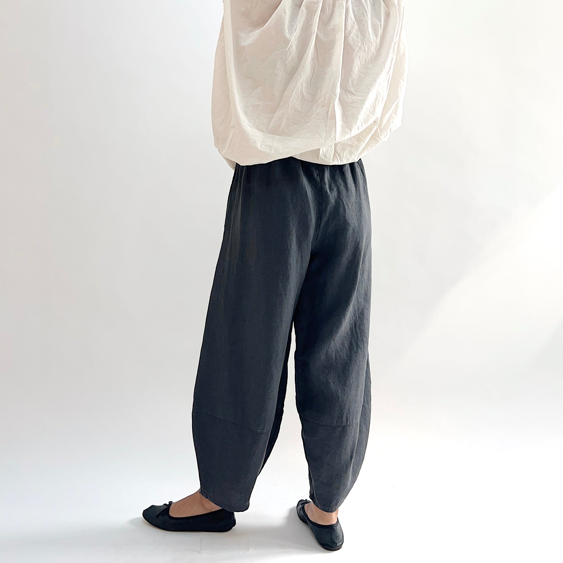Back view of model wearing the Bryn Walker Heavy Linen Oliver Pant in Moonlight, showing elastic waistband, full barrel-leg drape, and tapered ankle in deep moonlight charcoal heavy linen, styled with cream oversized top and black ballet flats
