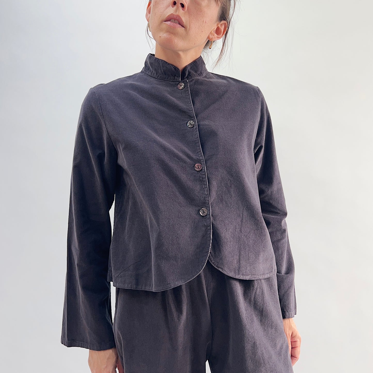 Model wearing the Bryn Walker Corduroy Standing Collar Crop Jacket in Fig, full front view showing standing collar, mother of pearl button closure, wide sleeves, curved crop hem, and deep fig pinwheel corduroy, styled with matching corduroy pants
