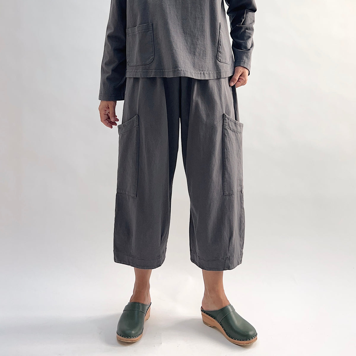 Model wearing the Bryn Walker Cotton Pasha Pant in Moonlight, full front view showing elastic gathered waistband, two large thigh patch pockets, wide-leg cropped length, and soft blue-grey cotton, styled with matching top and green wedge clogs
