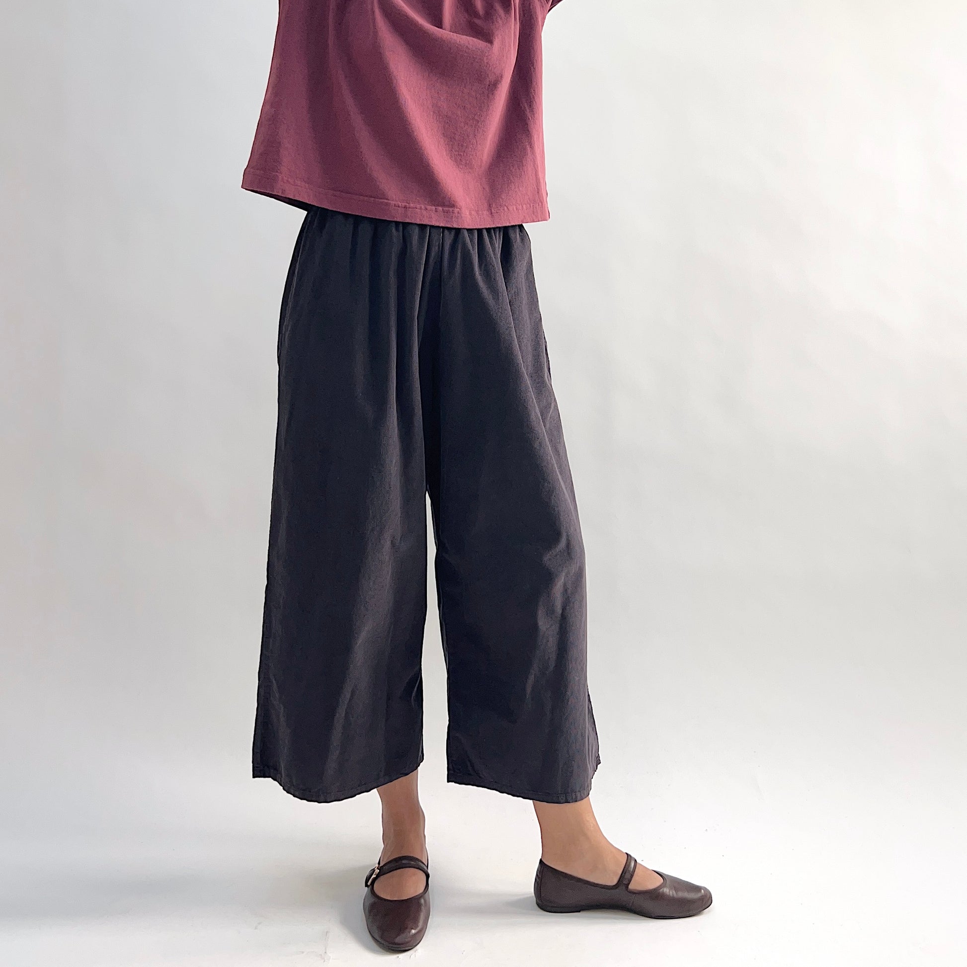 Model wearing the Bryn Walker Corduroy Flood Pants in Fig, front view showing fully elastic gathered waistband, wide culotte-style leg, mid-calf flood length, side pockets, and deep fig pinwheel corduroy, styled with burgundy Double Cotton Tee and brown Mary Jane flats
