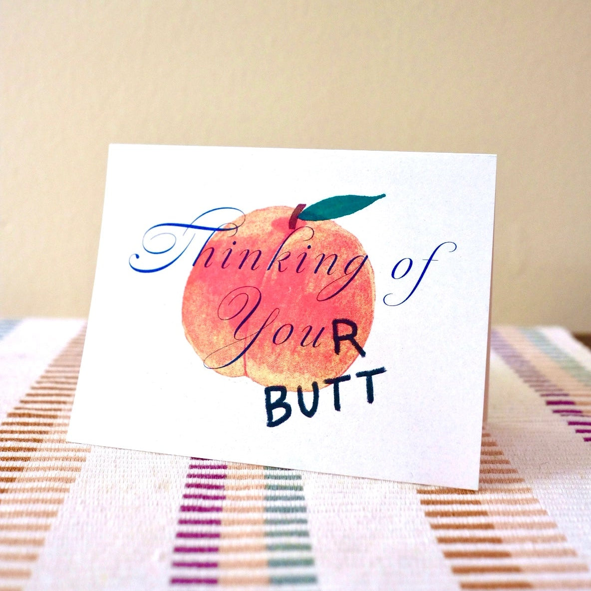 Small Adventure Thinking of Your Butt Card, illustrated peach with the words “Thinking of Your Butt,” displayed on striped fabric.
