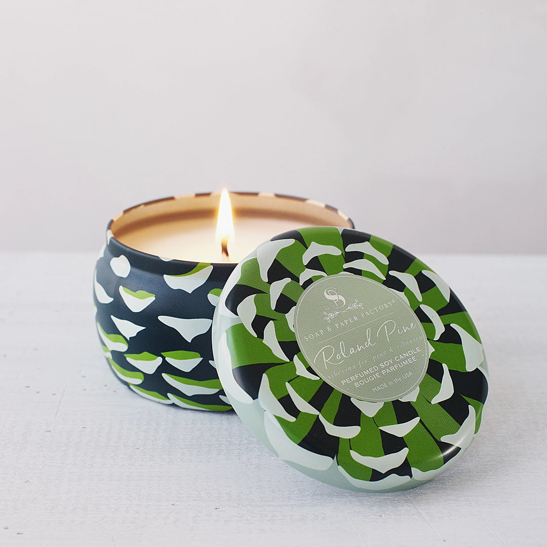 Lit Roland Pine large soy candle in a green and cream patterned tin, with the labeled lid resting beside it on white.

