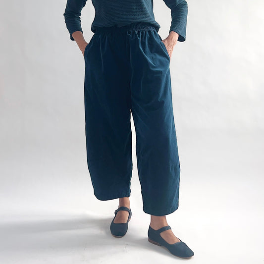 Full front view of model wearing deep teal mini cord lantern pants, hands in pockets, showing the gathered elastic waist, full lantern volume through the hip, and tapered ankle, styled with a matching teal textured long-sleeve top and teal mary jane flats against a white background.
