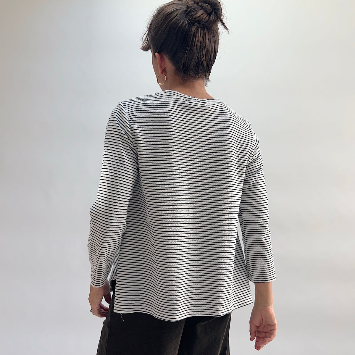 Cut Loose Striped Side Vent Crew in Laundered, white and black fine-stripe crewneck top showing side vent hem detail, hair in bun, paired with dark wide-leg trousers — back view
