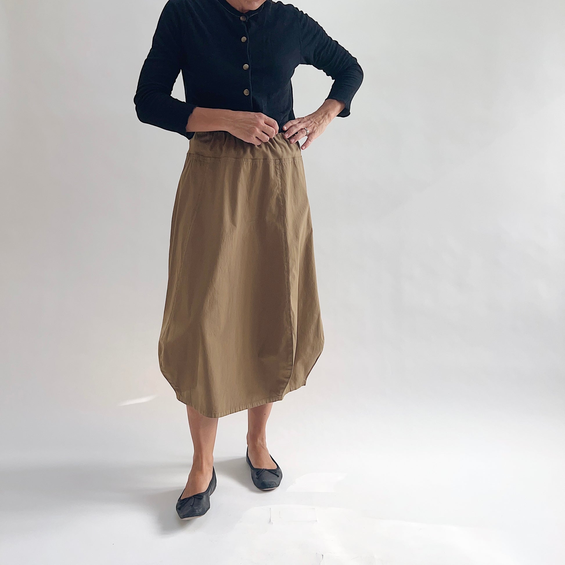 Full front view of model wearing Eleven Stitch winter olive rib yoke skirt with hands at waistband, showing elastic knit waistband, curved seam paneling, sculptural bubble hem, below-the-knee length, and warm khaki olive color, styled with black button-front cardigan and black ballet flats