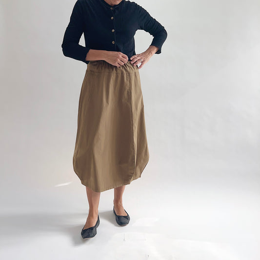Full front view of model wearing Eleven Stitch winter olive rib yoke skirt with hands at waistband, showing elastic knit waistband, curved seam paneling, sculptural bubble hem, below-the-knee length, and warm khaki olive color, styled with black button-front cardigan and black ballet flats