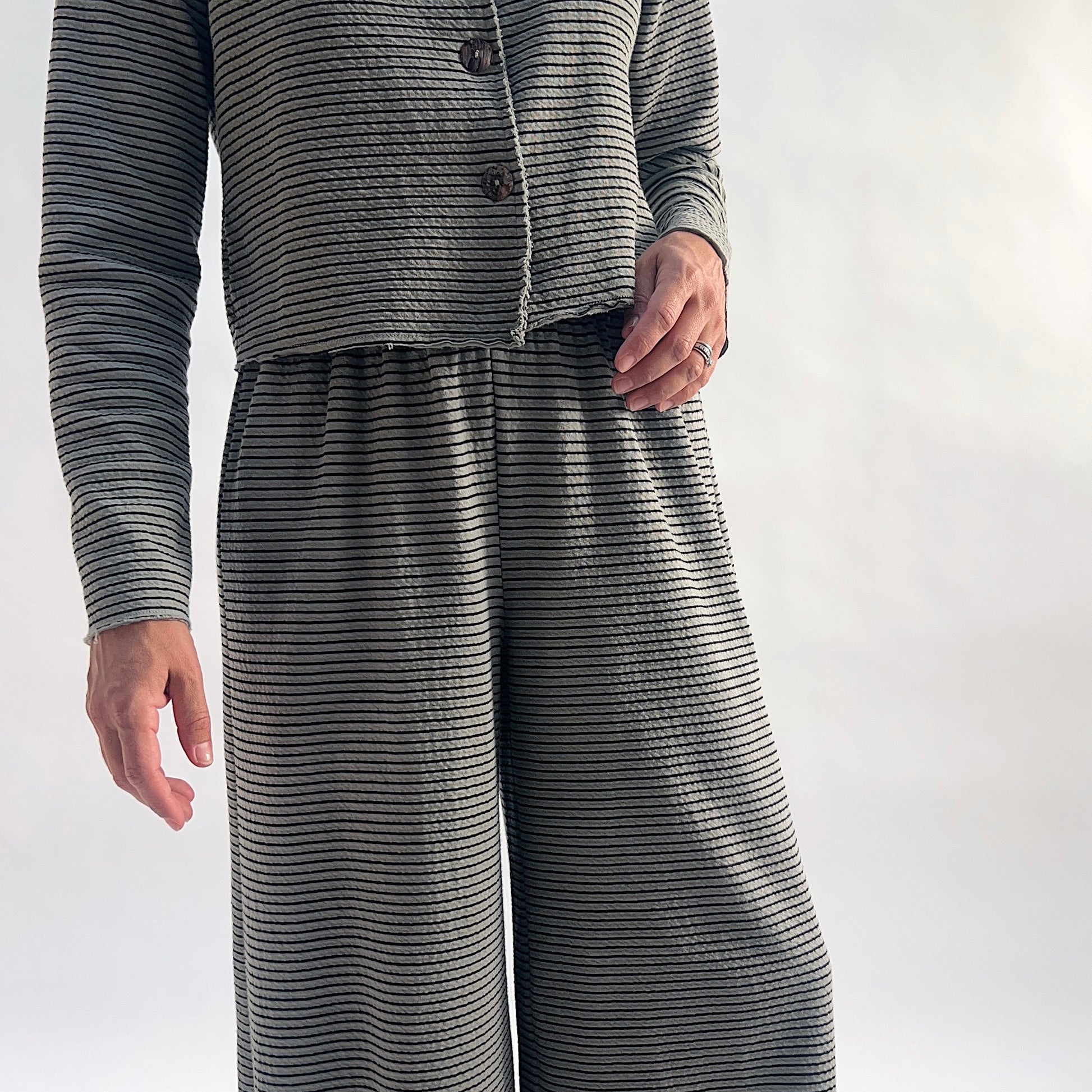 Close crop of model wearing squirrel and black fine horizontal stripe wide-leg cropped pants with dart shaping, showing the elastic waist and crinkle-texture fabric, styled with the matching striped asymmetrical cardigan against a white background.
