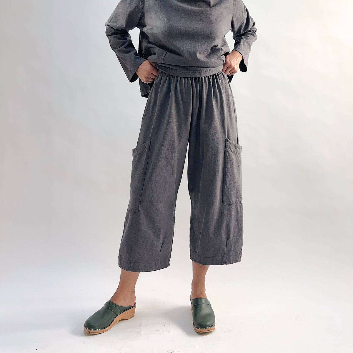 Model with hands on hips wearing the Bryn Walker Cotton Pasha Pant in Moonlight, front view showing fully elastic gathered waistband, two large thigh patch pockets, wide-leg cropped silhouette, and soft blue-grey cotton, styled with matching top and green wedge clogs
