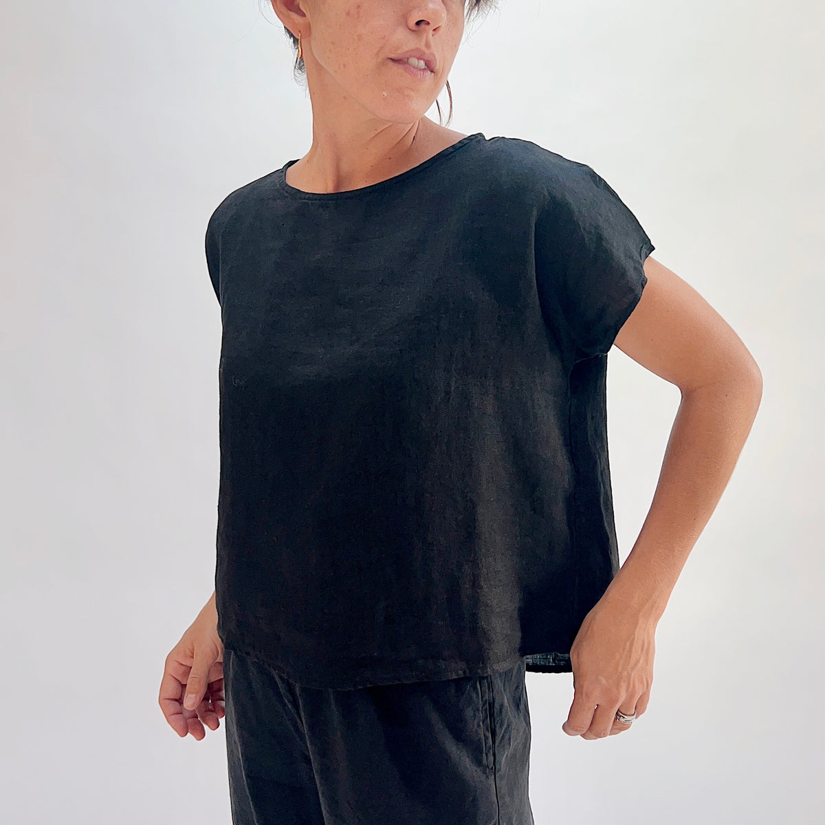 Front close-up of black 100% linen high-low tee with boatneck neckline and cap sleeves, showing relaxed fit and woven linen texture, hand on hip, paired with black linen lantern pants
