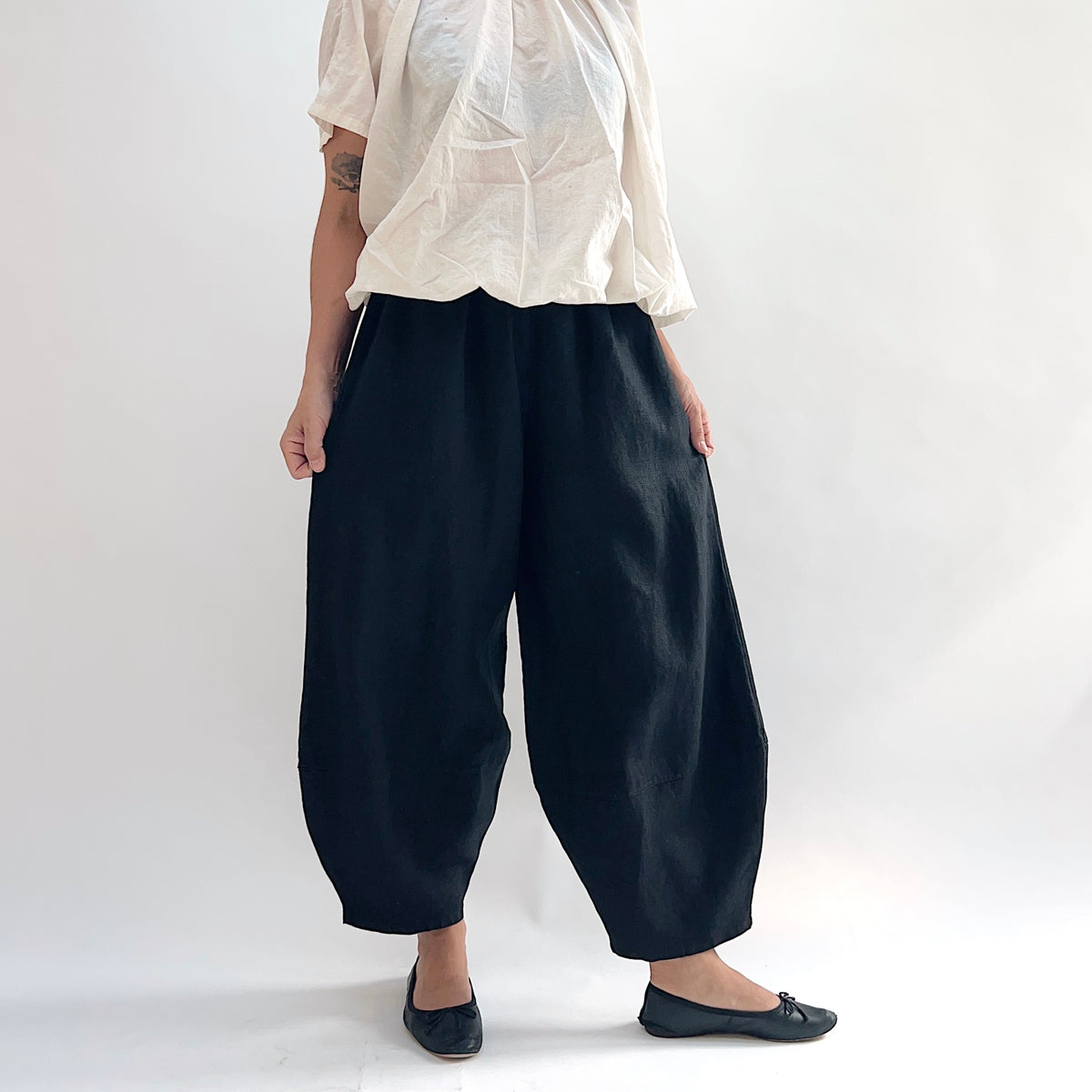 Model wearing the Bryn Walker Heavy Linen Oliver Pant in Black, front view showing barrel-leg silhouette, elastic waistband, side pockets, and tapered ankle in black heavy linen, styled with cream short-sleeve linen top and black ballet flats
