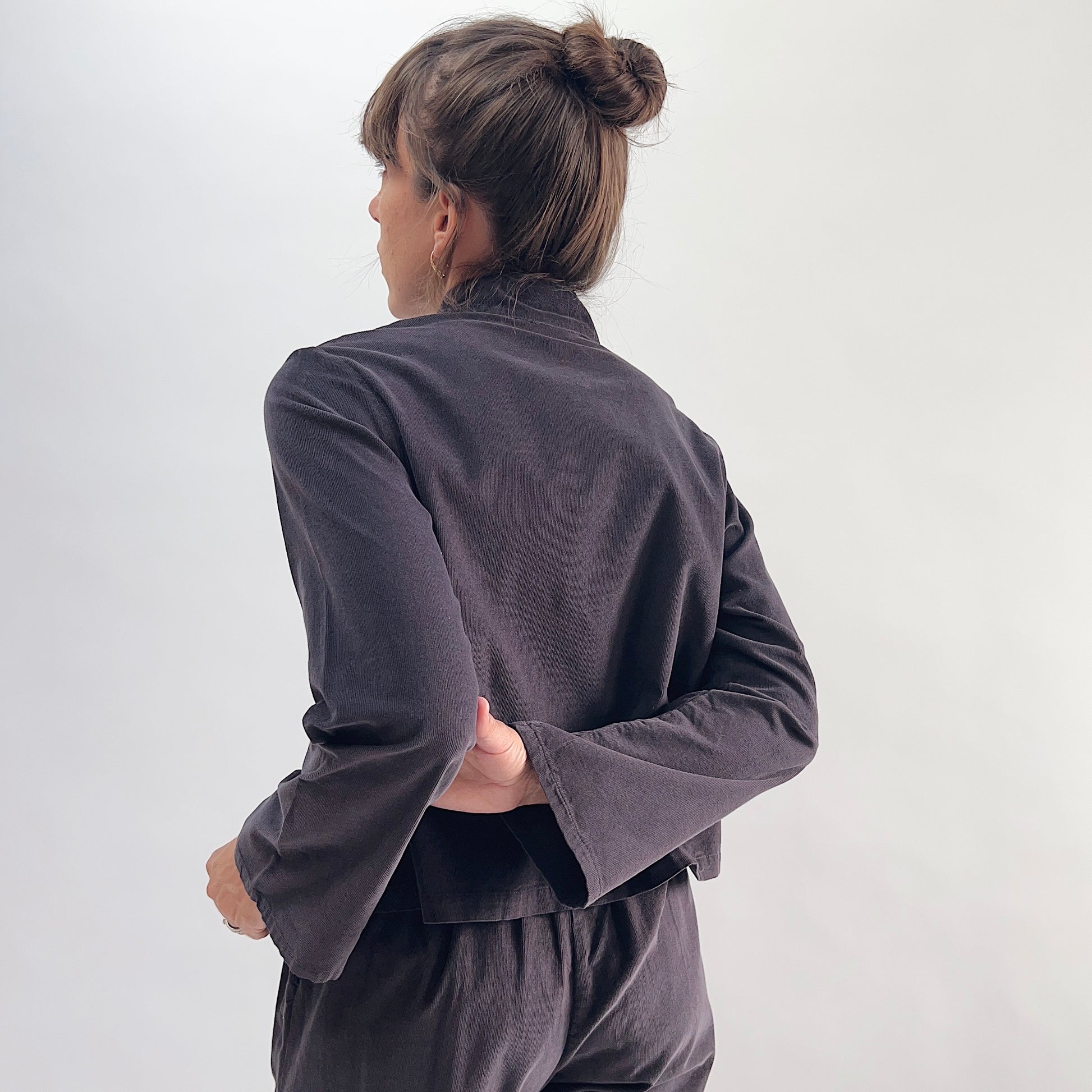 Back view of model wearing the Bryn Walker Corduroy Standing Collar Crop Jacket in Fig, showing standing collar, wide sleeves with slit cuff detail, curved crop hem, and deep fig pinwheel corduroy texture, styled with matching corduroy pants
