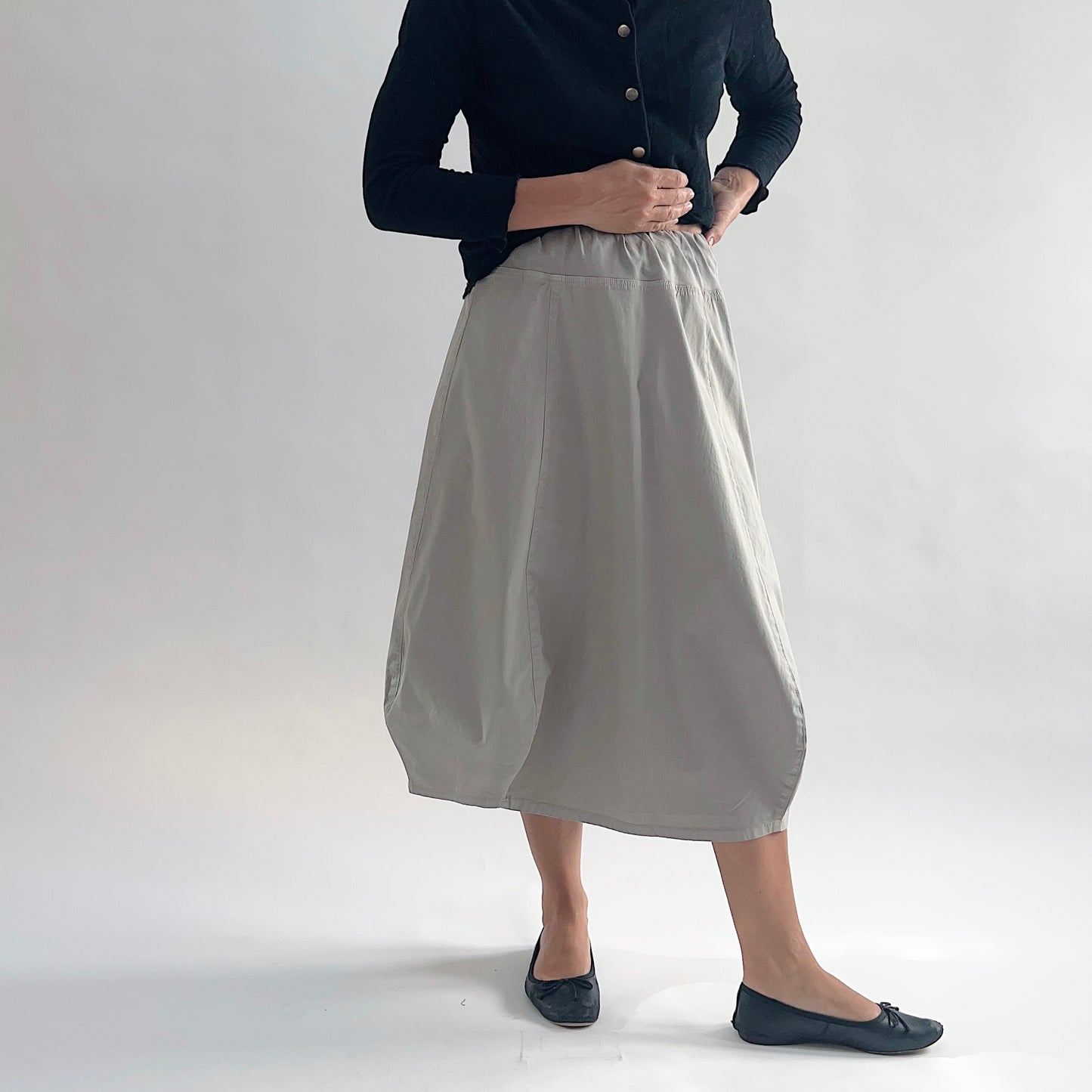 Full front view of model wearing Eleven Stitch fennel rib yoke skirt with hands on hips, showing elastic knit waistband, curved seam paneling, sculptural bubble hem, below-the-knee length, and warm taupe color, styled with black button-front cardigan and black ballet flats