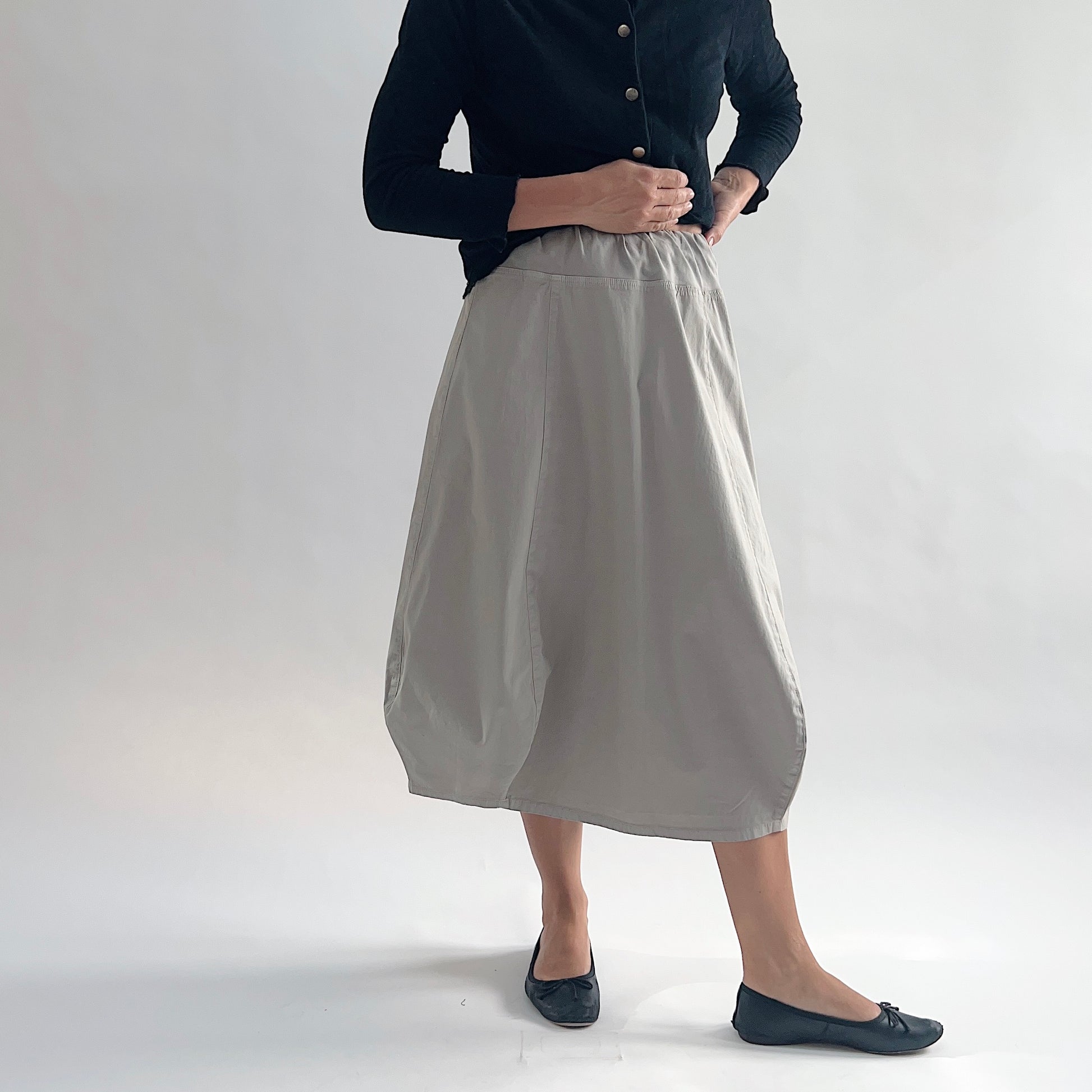 Full front view of model wearing Eleven Stitch fennel rib yoke skirt with hands on hips, showing elastic knit waistband, curved seam paneling, sculptural bubble hem, below-the-knee length, and warm taupe color, styled with black button-front cardigan and black ballet flats