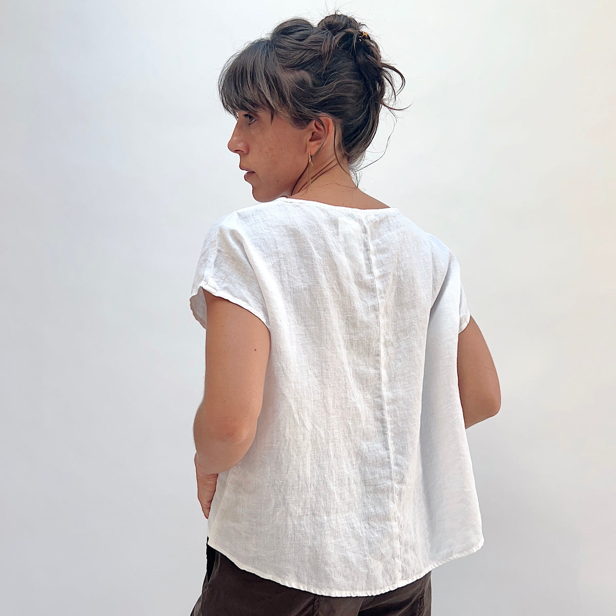 Back view of white 100% linen high-low tee with boatneck, cap sleeves, and center back seam detail, worn with dark trousers, model's hair up
