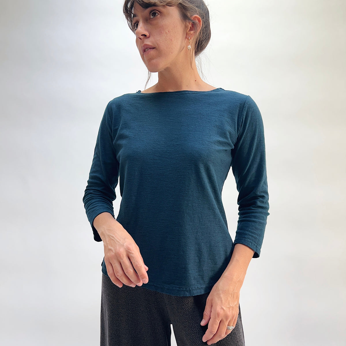Model wearing the Cut Loose 3/4 Sleeve Boatneck Top in Etan, front view looking up, deep teal cotton-linen boatneck top with wide neckline, 3/4 sleeves, and relaxed fit, styled with black textured pants
