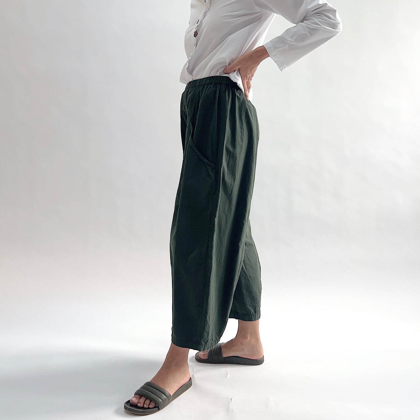 Side view of model wearing Eleven Stitch konbu one pocket pant with hand on hip, showing elastic waistband, wide curved-seam cropped leg, large side pocket, gathered elastic hem, and deep forest green color, styled with white long sleeve button front shirt and dark slide sandals