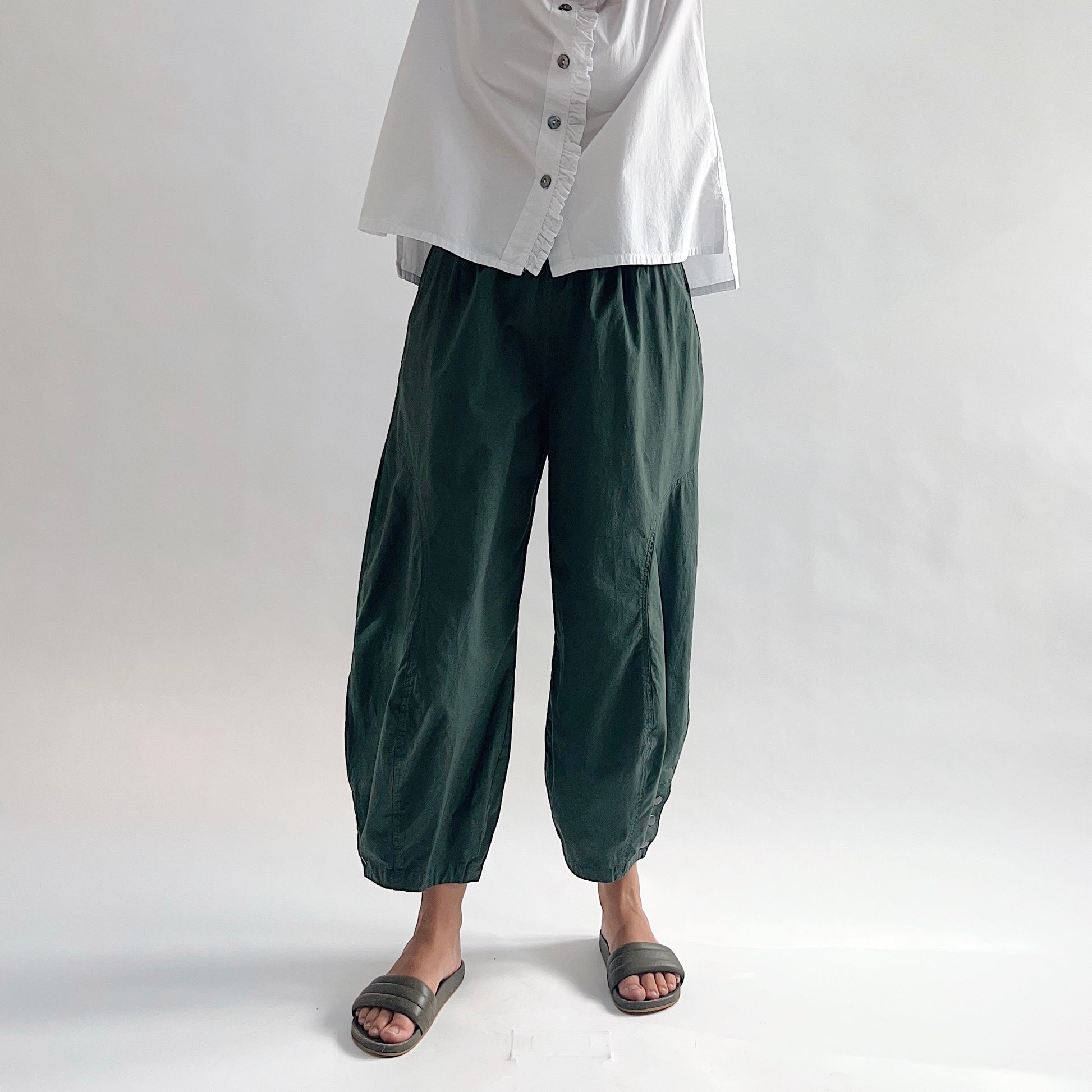 Full front view of model wearing Eleven Stitch konbu snap doreen pant showing elastic waistband, wide balloon leg tapering to snap hem at ankle, snap buttons at ankle, and deep forest green color, styled with white blossom collar shirt and dark slide sandals